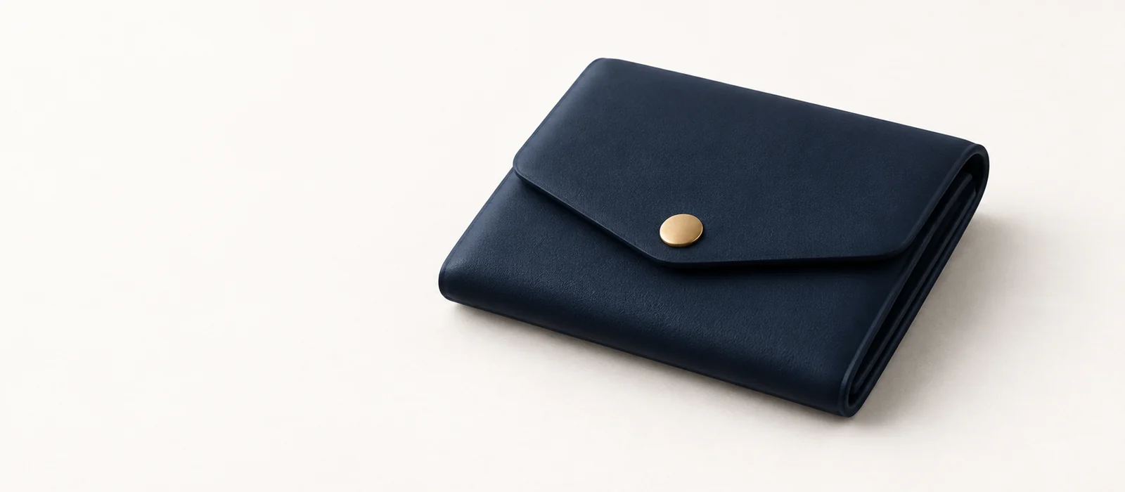 A closed matte navy wallet with a small gold snap, on warm paper.