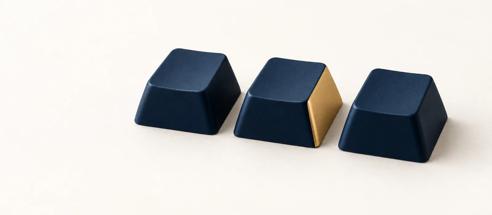 Three blank matte navy keycaps in a row, one with a gold side, on warm paper.